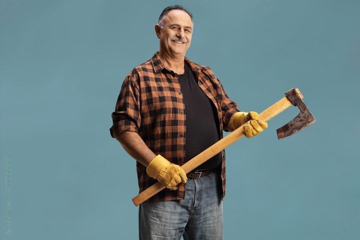 A smiling man in wearing checkered polo, black shirt and yellow gloves is holding a rusty axe.