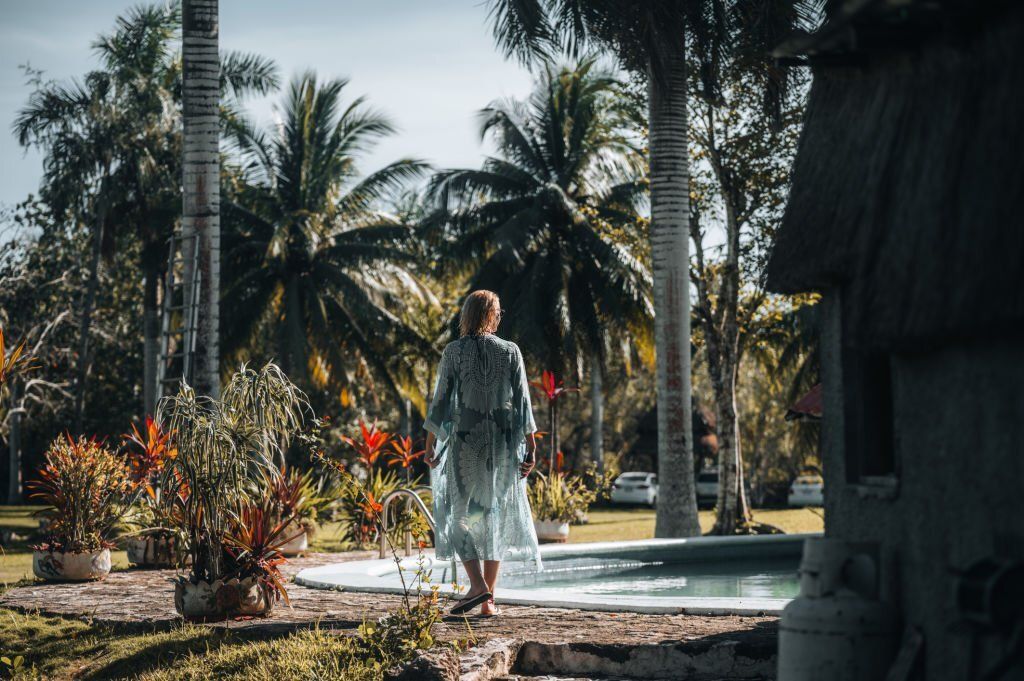 Woman stands by the poolside surrounded by palm trees and tropical plants.