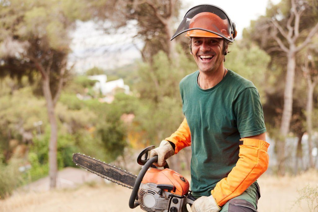 An arborist wearing a hardhat with face guard, green shirt, and orange armguard is smiling while holding a chainsaw.