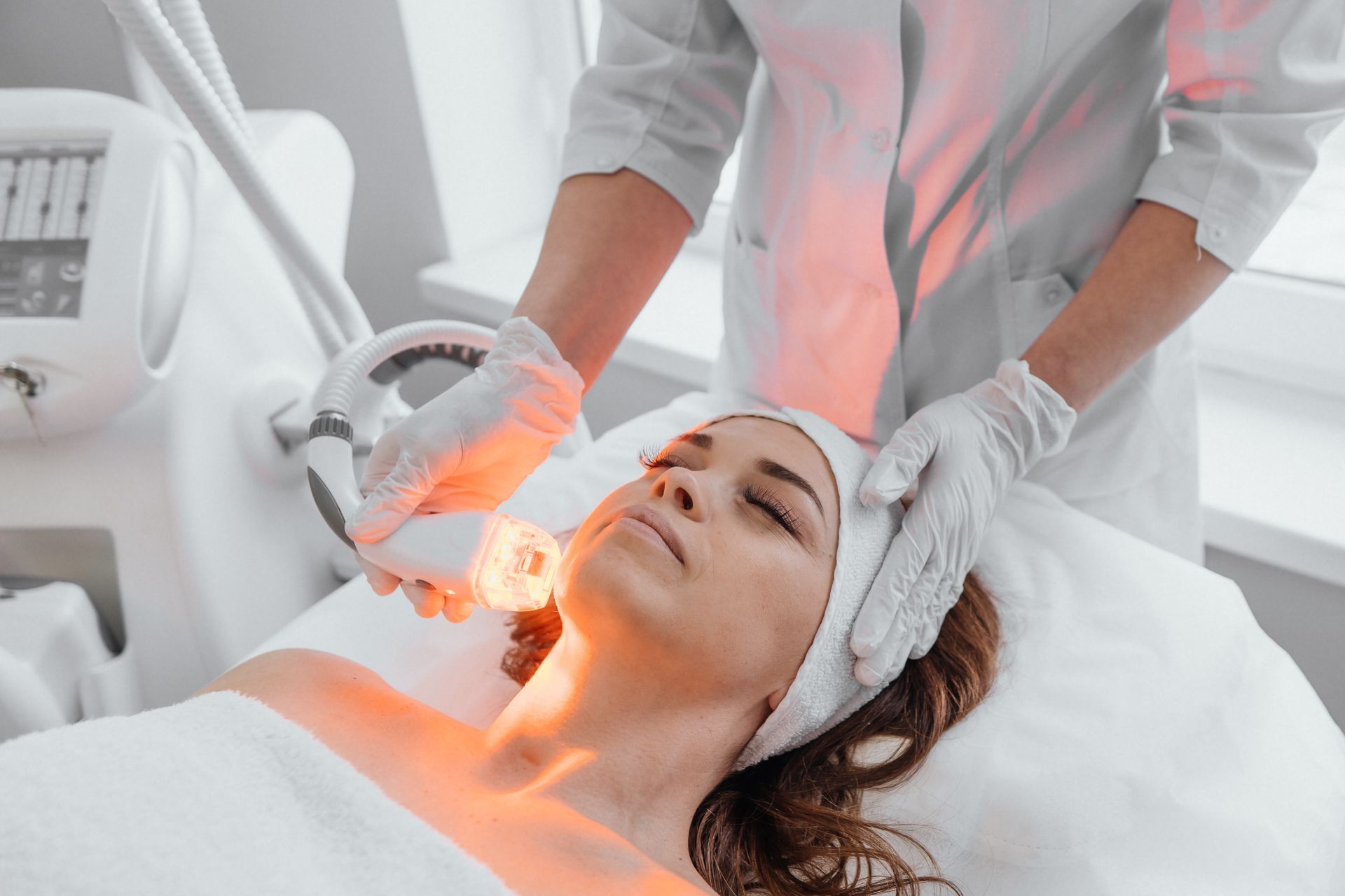 Woman receiving light therapy facial treatment; red light, white room, technician.