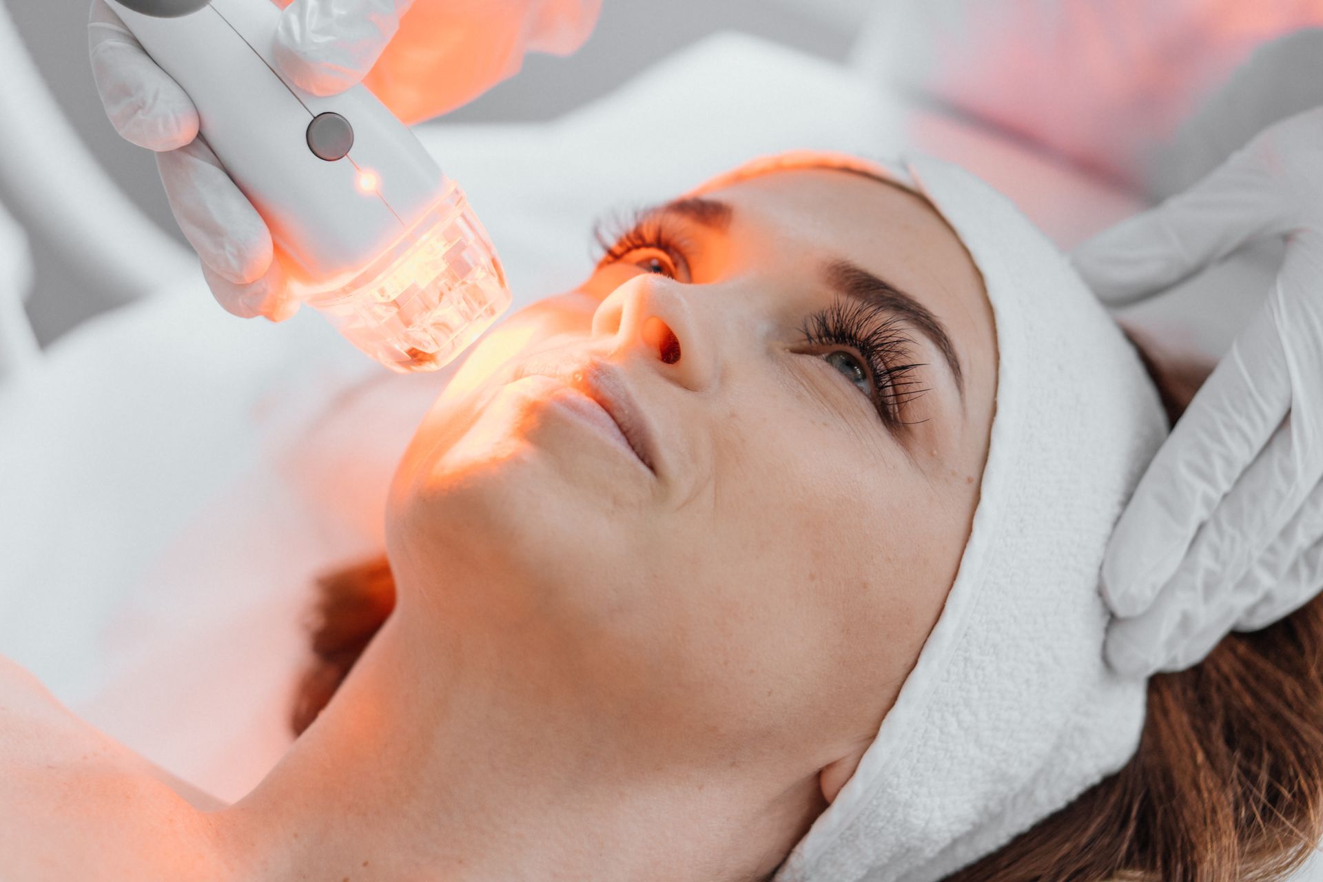 Woman receiving facial treatment with a light device, hands in gloves, warm light.