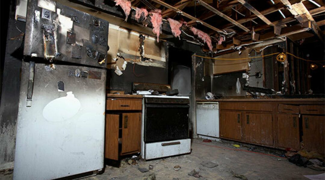 A kitchen interior heavily damaged by fire, featuring charred cabinets, a blackened ceiling, and exposed insulation.
