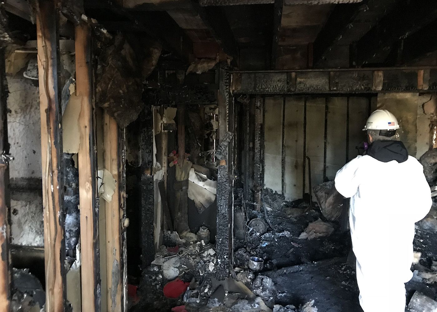 A person in a white hazmat suit and hard hat surveys the interior of a fire-damaged building with charred wooden framing.
