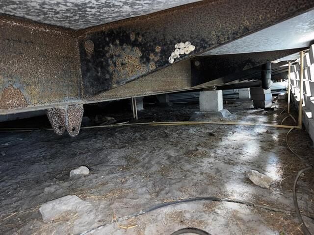 The underside of a mobile home structure featuring a metal frame, concrete piers, and ground covered by a vapor barrier.