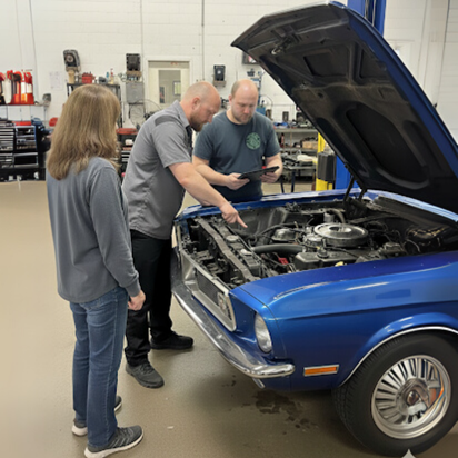 Three people examining a blue car's engine in a shop. Two men point; the woman looks on.