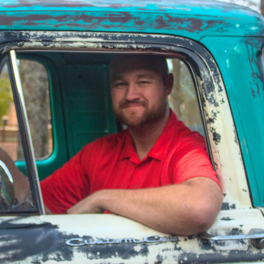 Man in red shirt smiles, leans out of teal and white vintage truck window.