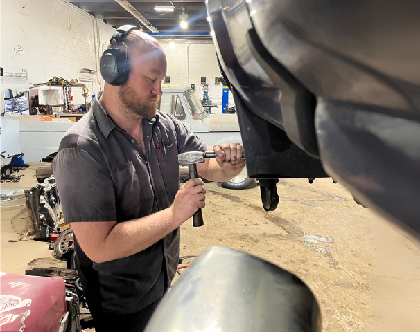 Mechanic hammering on a car fender in a garage. Wearing ear protection, focused.