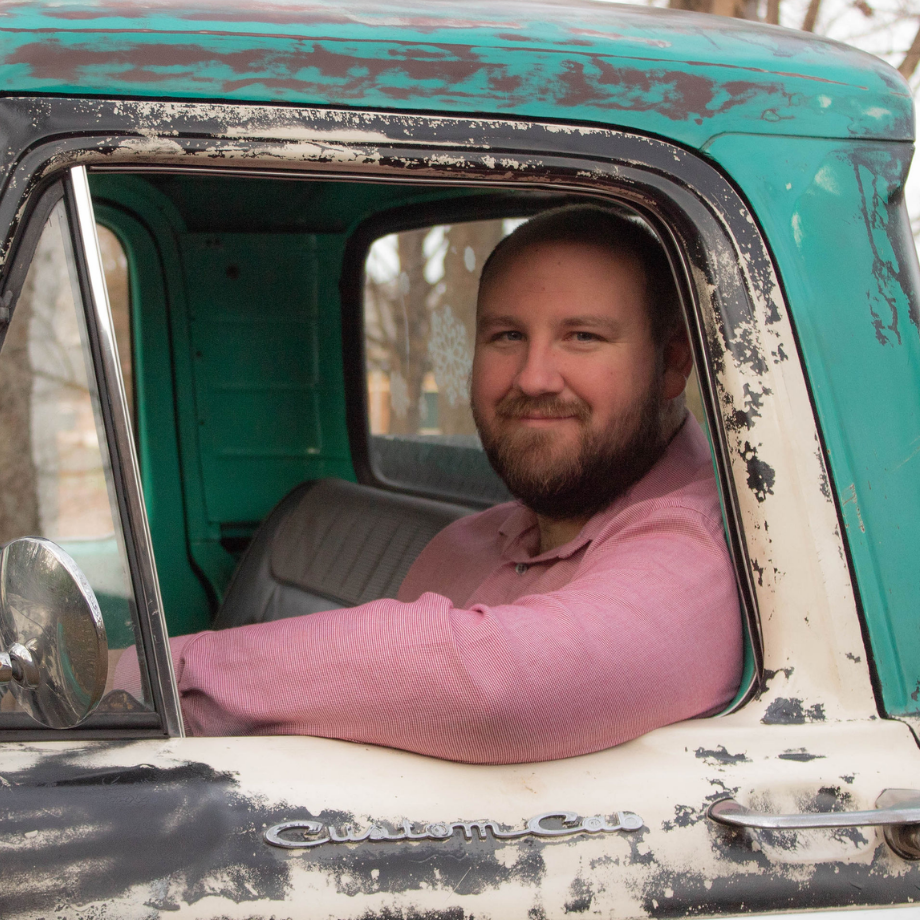 Man in a pink shirt smiles, sitting in the driver's seat of a weathered, teal and white truck.