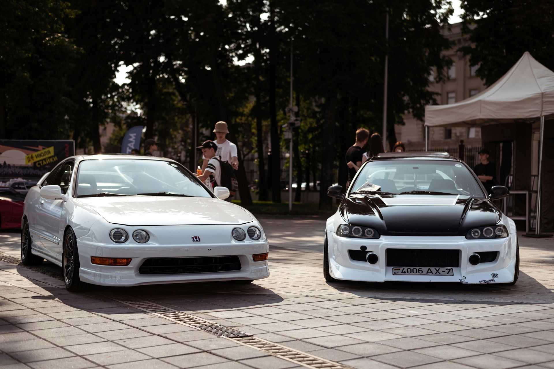 Two modified cars, white and black, parked on cobblestones with people and tents in background.