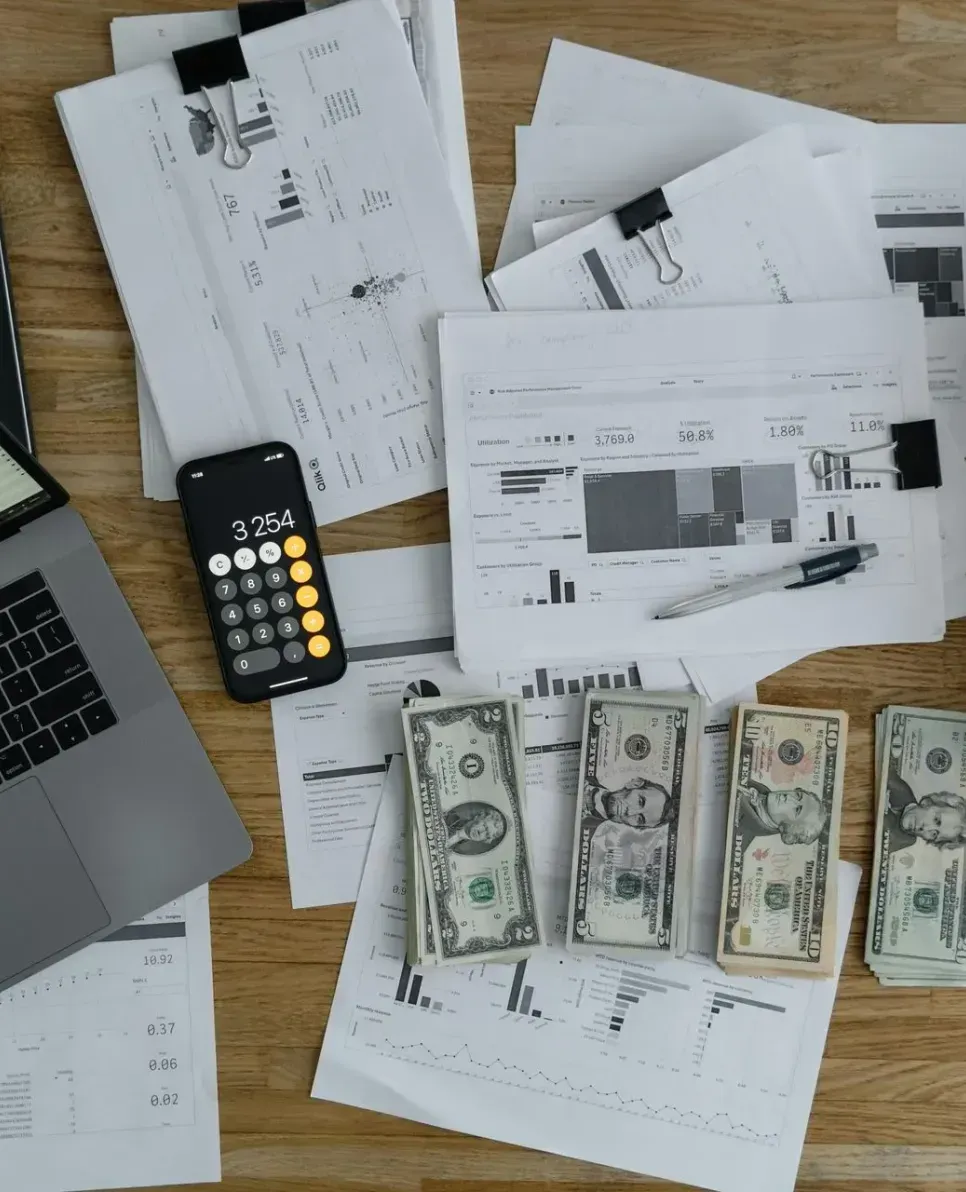 A wooden desk with a laptop , calculator , and money on it.