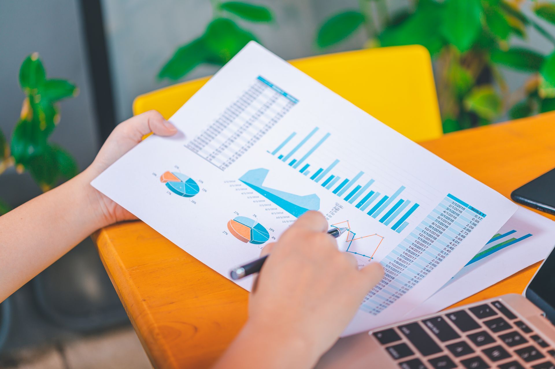 Person reviews financial reports with charts and graphs, using a pen, near a laptop and plants.