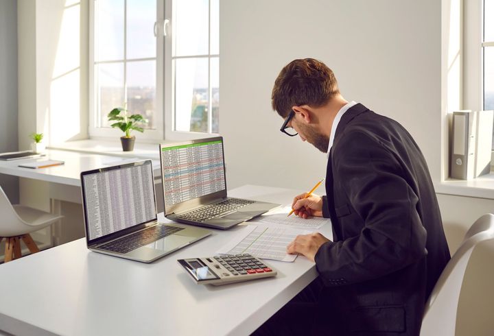 A man is sitting at a desk with two laptops and a calculator.