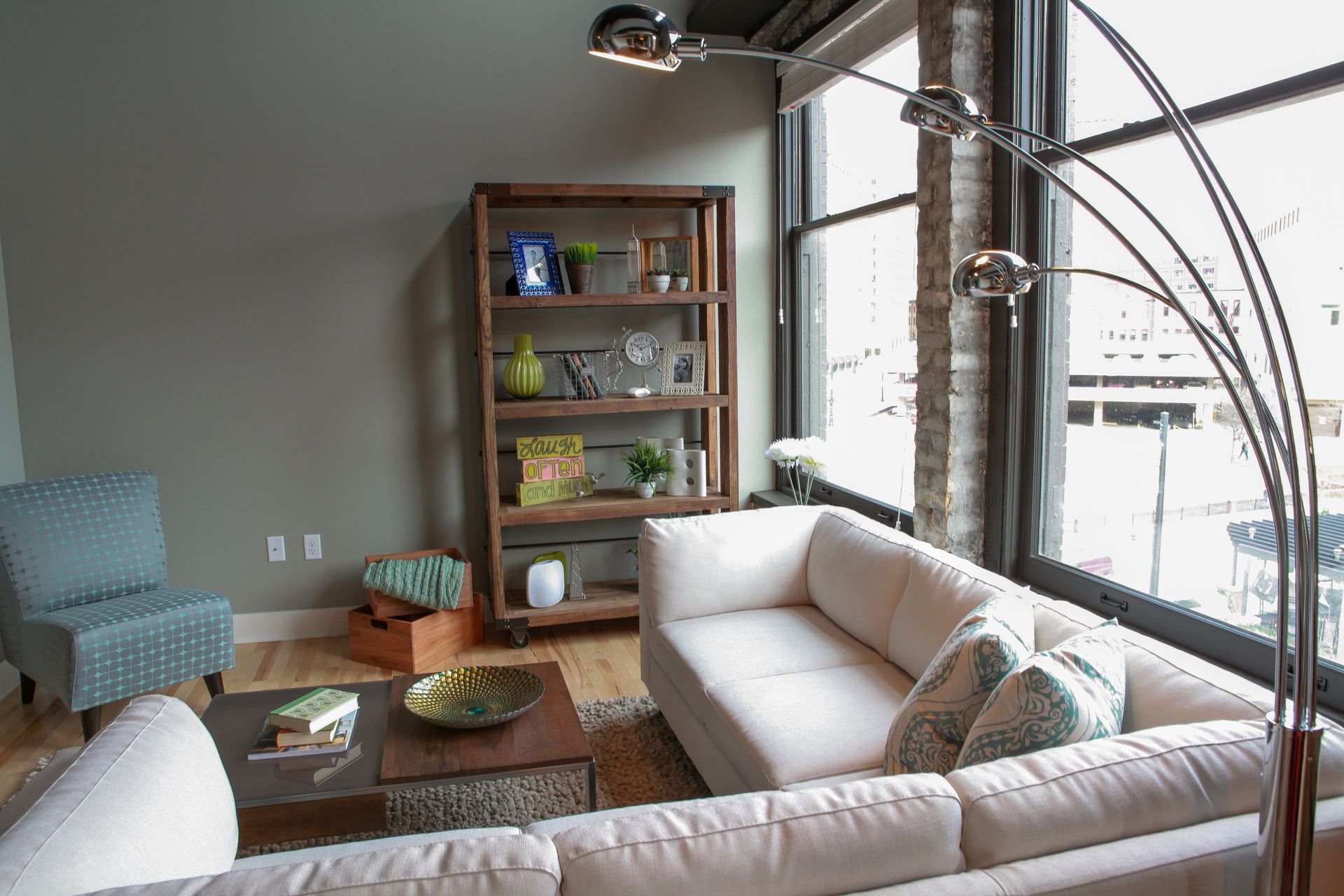 Bright living room with white sectional sofa, coffee table, armchair, bookshelf, and large windows.