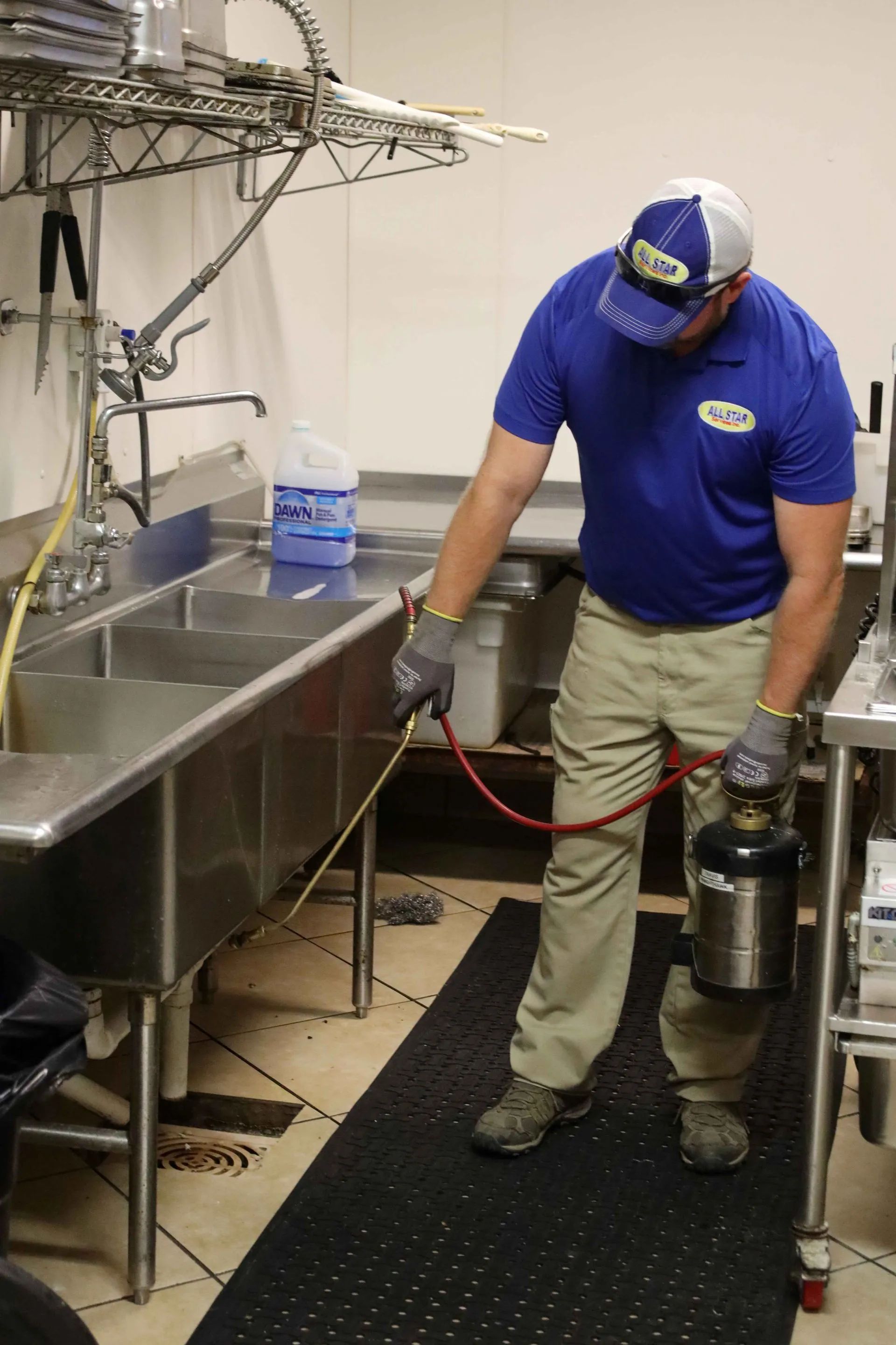 A technician in a blue uniform sprays liquid from a pressurized tank along the base of a stainless steel kitchen sink.