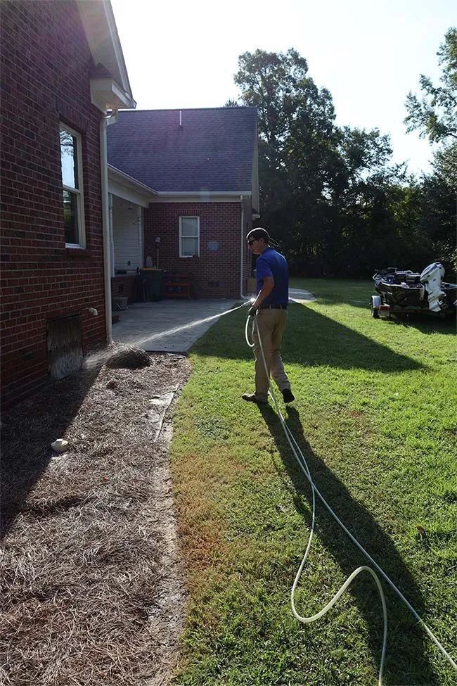 A person in a blue shirt and khaki pants uses a hose to spray water near the brick wall of a house.