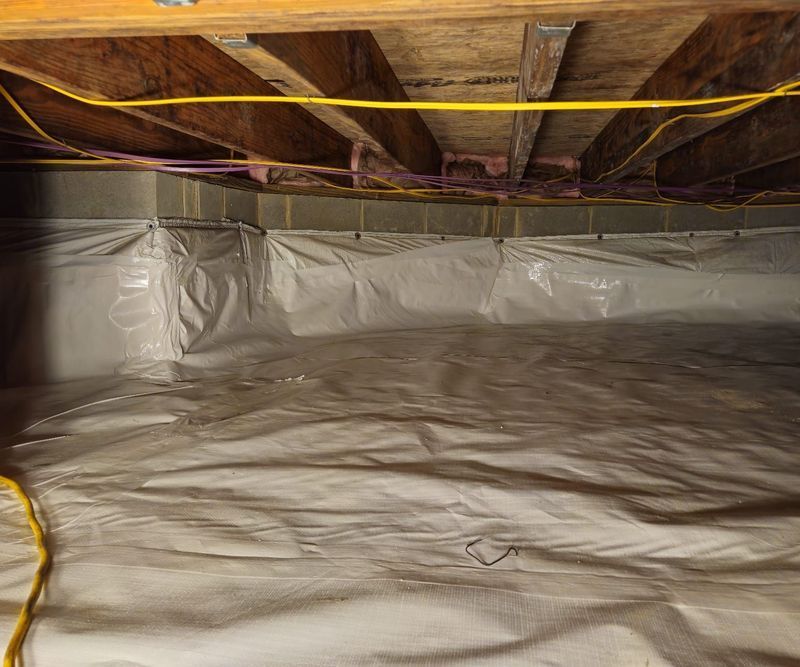 Crawl space with a white vapor barrier covering the ground and walls, featuring wooden floor joists and electrical cables.