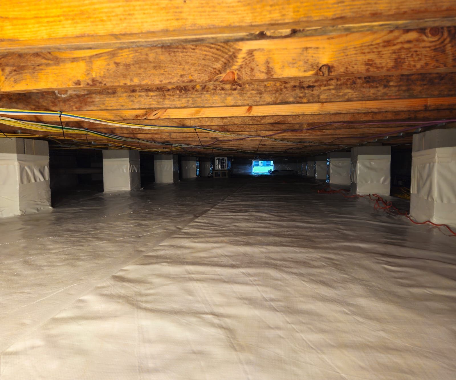 A crawl space with a white vapor barrier on the ground and wrapped concrete piers supporting wooden floor joists above.