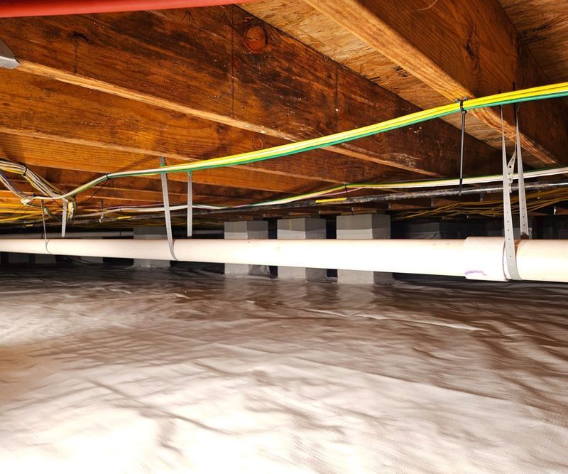 A white pipe suspended by metal straps from wooden floor joists above a white vapor barrier in a crawl space.