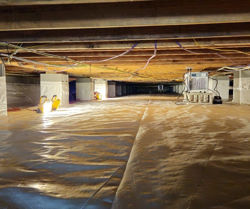 A crawl space with a vapor barrier on the ground, wrapped foundation piers, and a dehumidifier unit in the background.