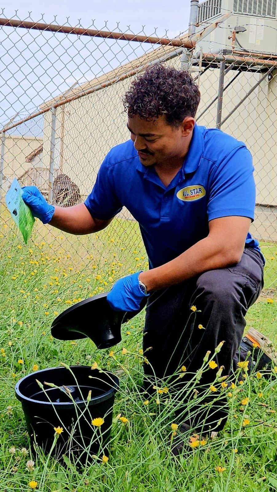 A person in a blue polo shirt and gloves kneels in a grassy field to place a black bucket near a chain-link fence.