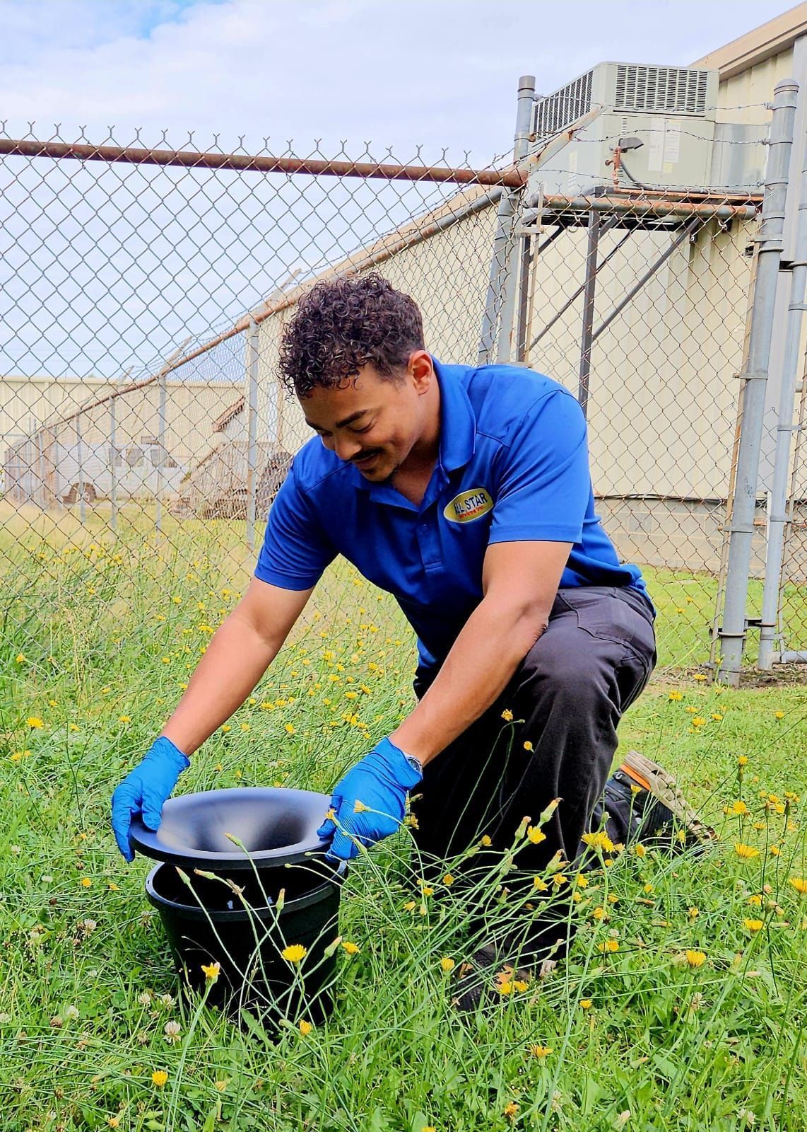 A person in a blue shirt and blue gloves kneels in grass, setting a black funnel trap near a chain-link fence.