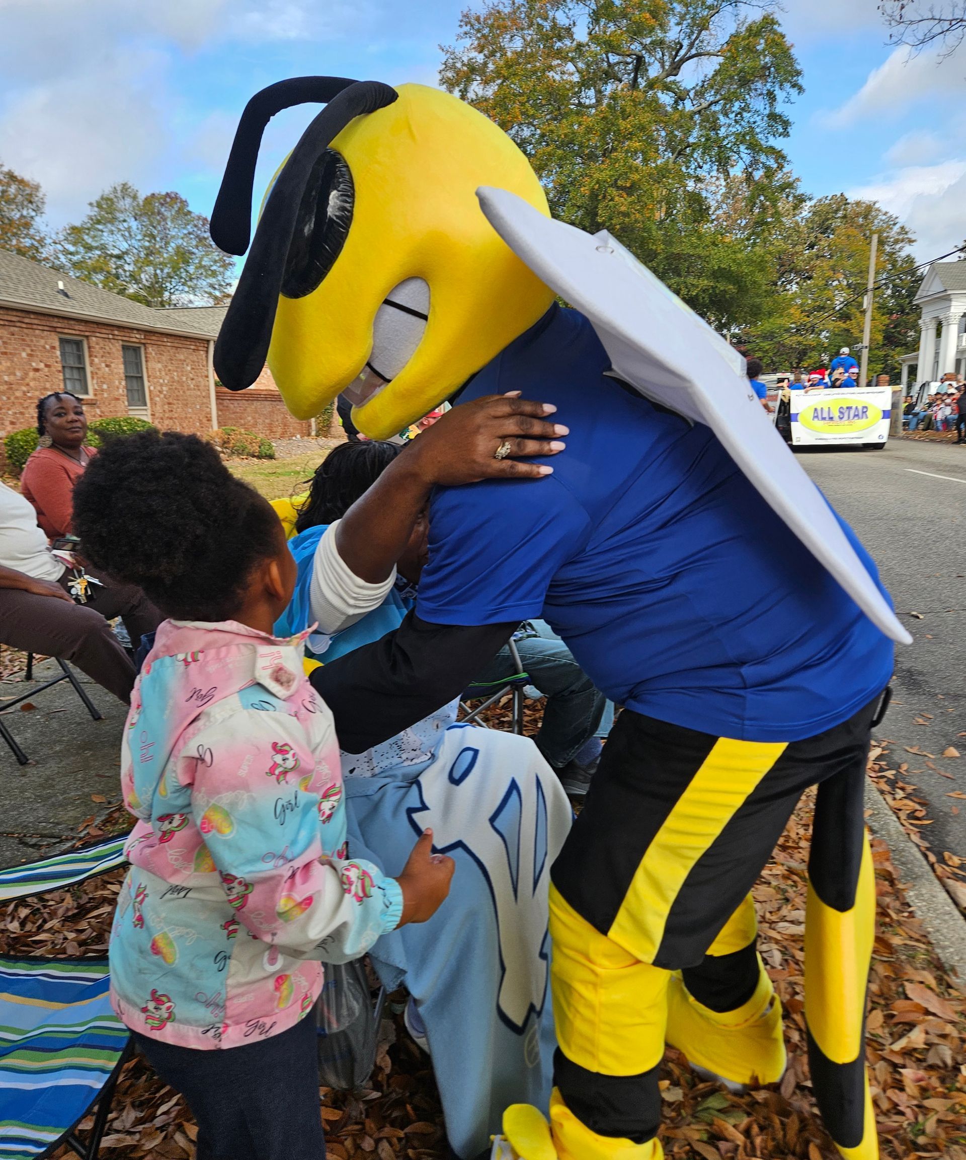 A mascot dressed as a yellow jacket bee hugs a person while a child looks on during an outdoor parade.