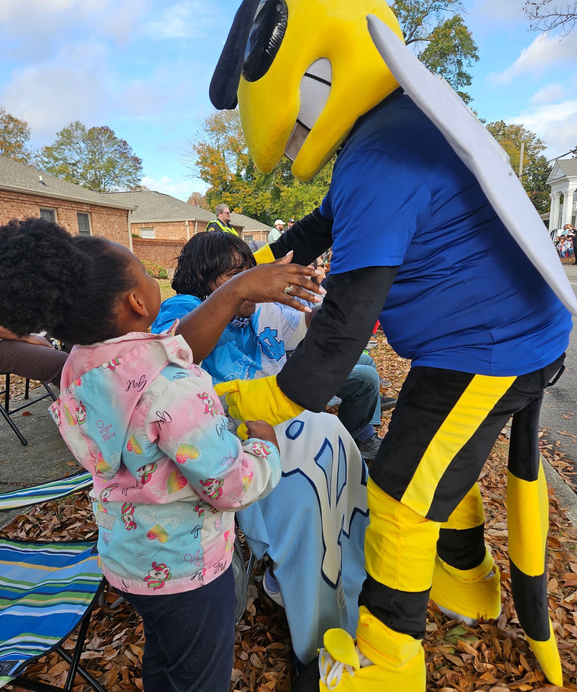 A yellow-and-black insect mascot interacts with a child and another person outdoors on a sunny day.