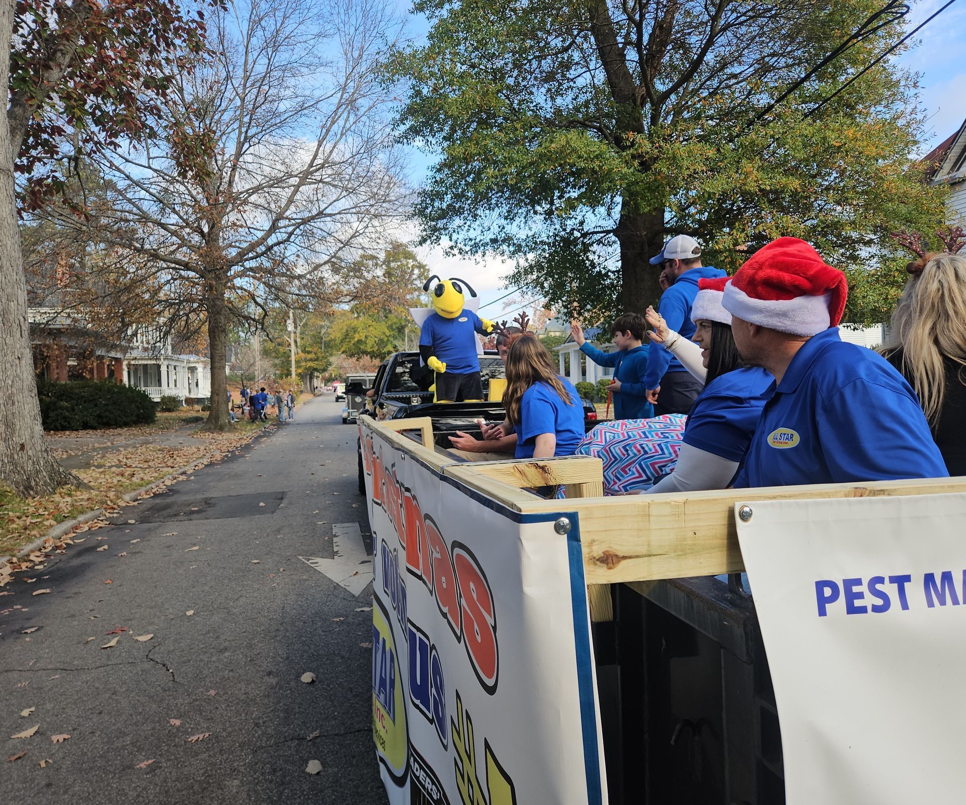 A parade float with a wasp mascot, people in blue shirts and Santa hats, moving down a street on a sunny autumn day.
