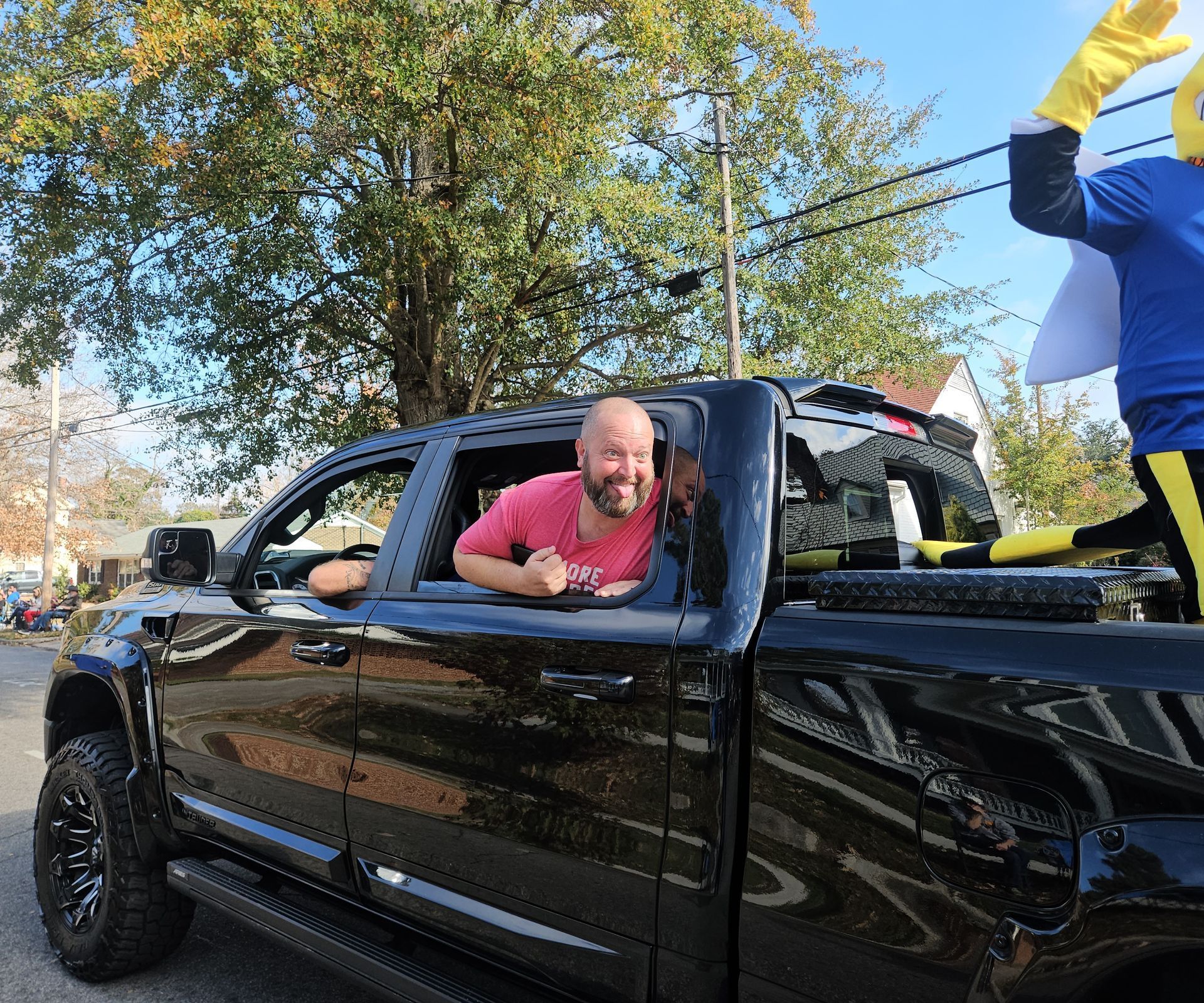 A smiling person leans out the window of a black pickup truck, while a costumed mascot waves nearby under sunny trees.