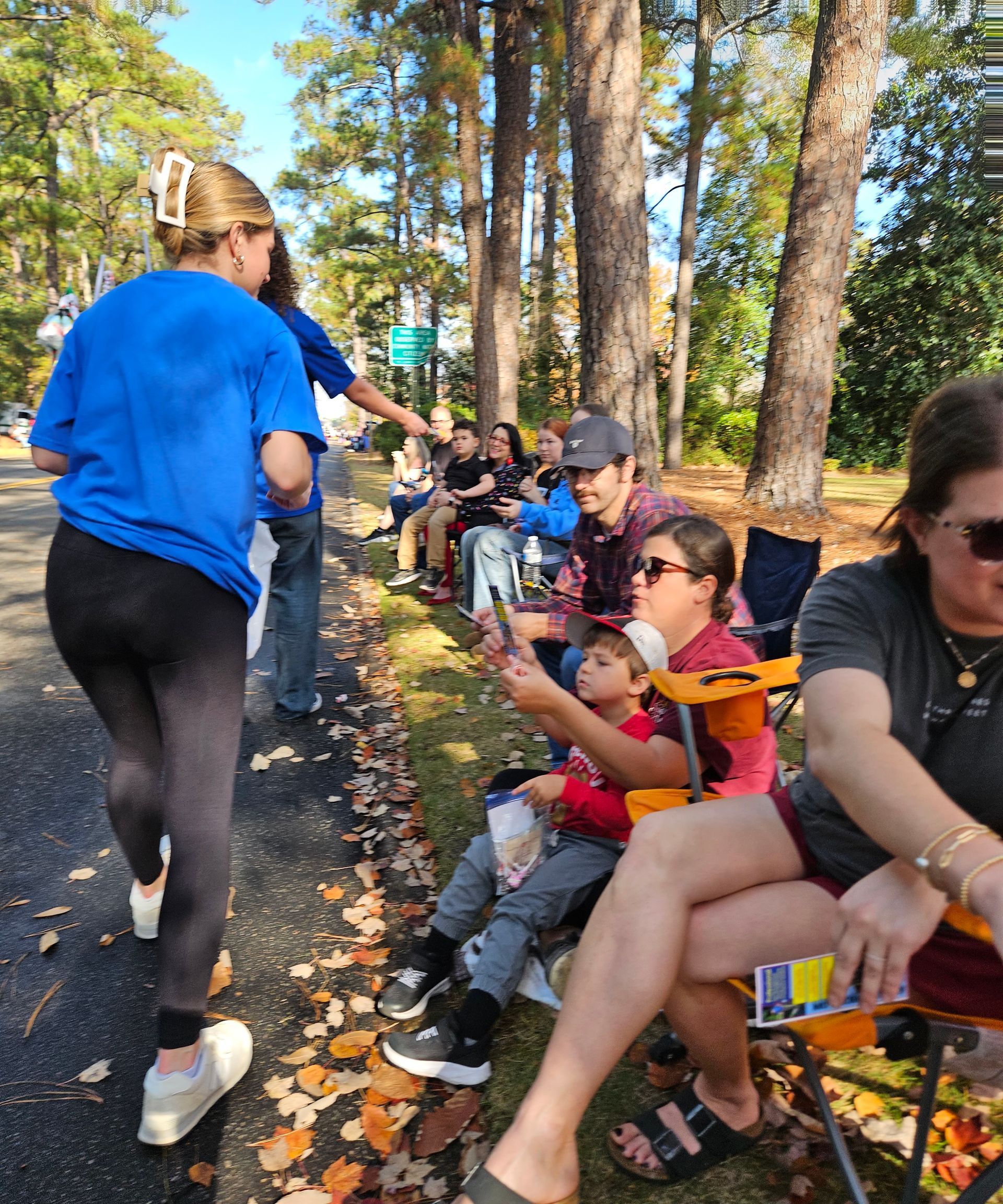 People in casual clothing sit on chairs along a road, some holding items, while someone walks by in the foreground.