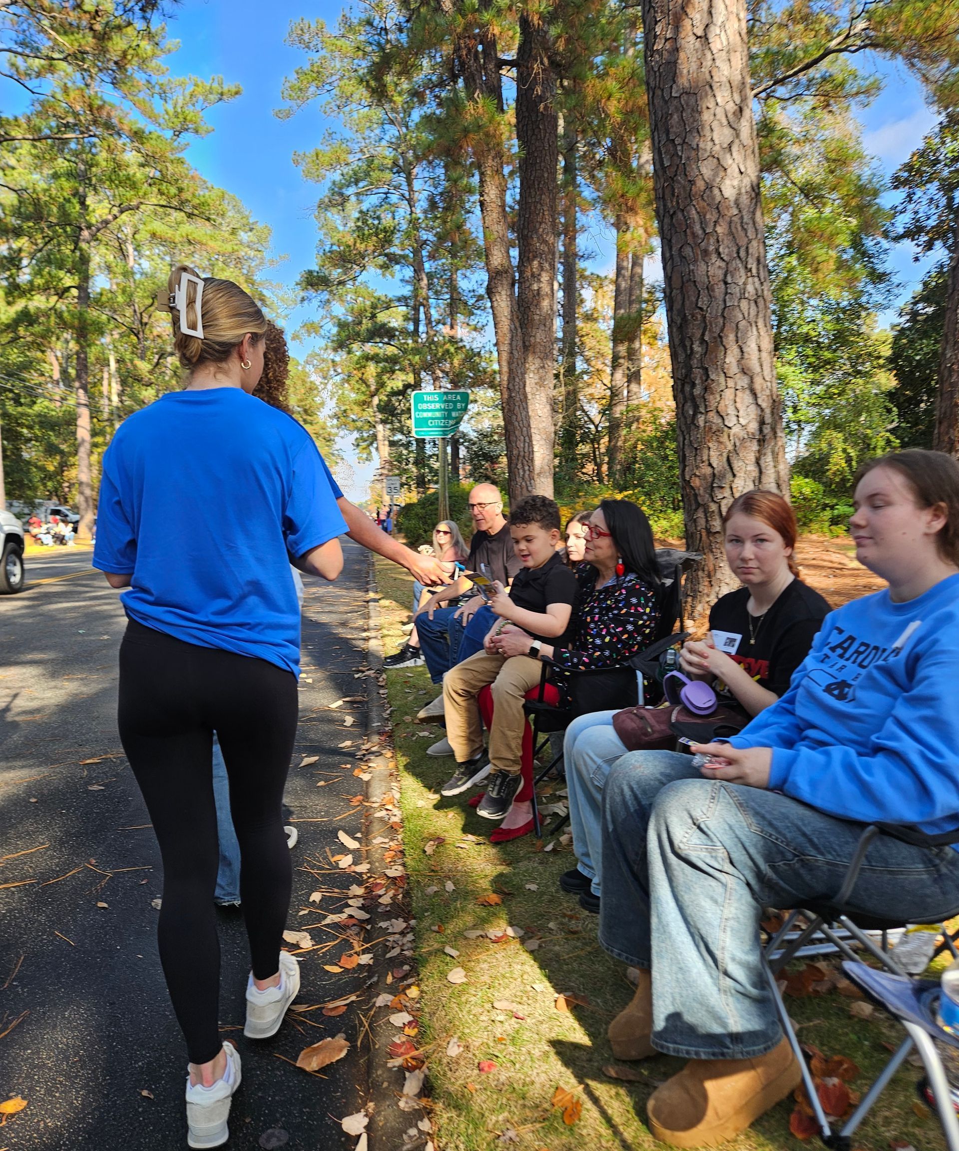 A line of people sitting on portable chairs along a road, with one person standing and reaching out to them.