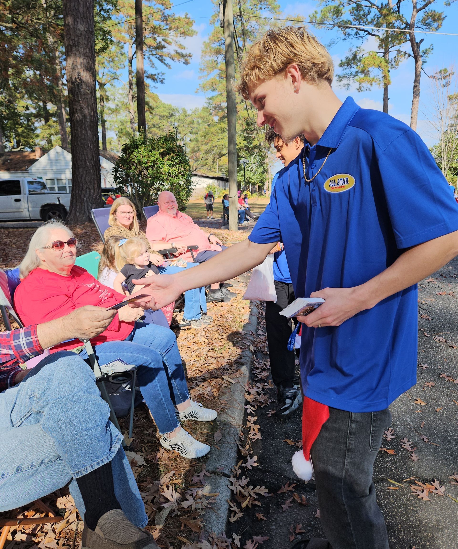 A young person in a blue polo shirt hands a small item to a seated person outdoors, with others watching in the background.