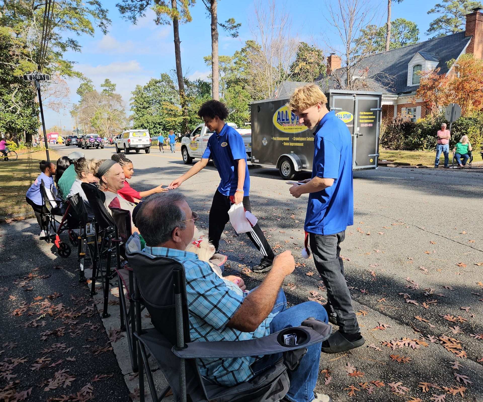 Two people in blue shirts hand items to seated onlookers along a gravel road on a sunny day.