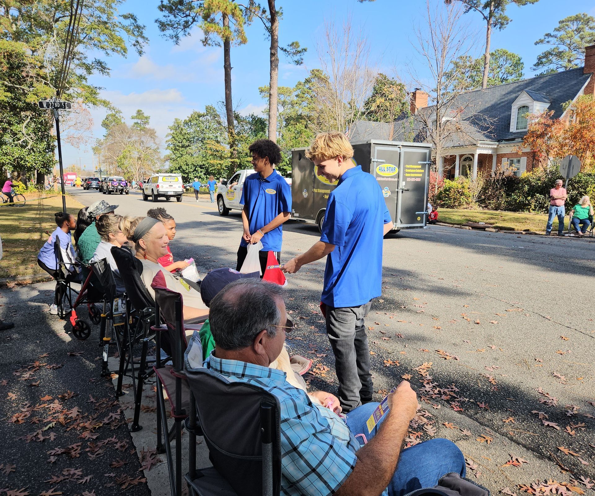 Two people in matching blue shirts hand items to seated onlookers along a sunny residential street.