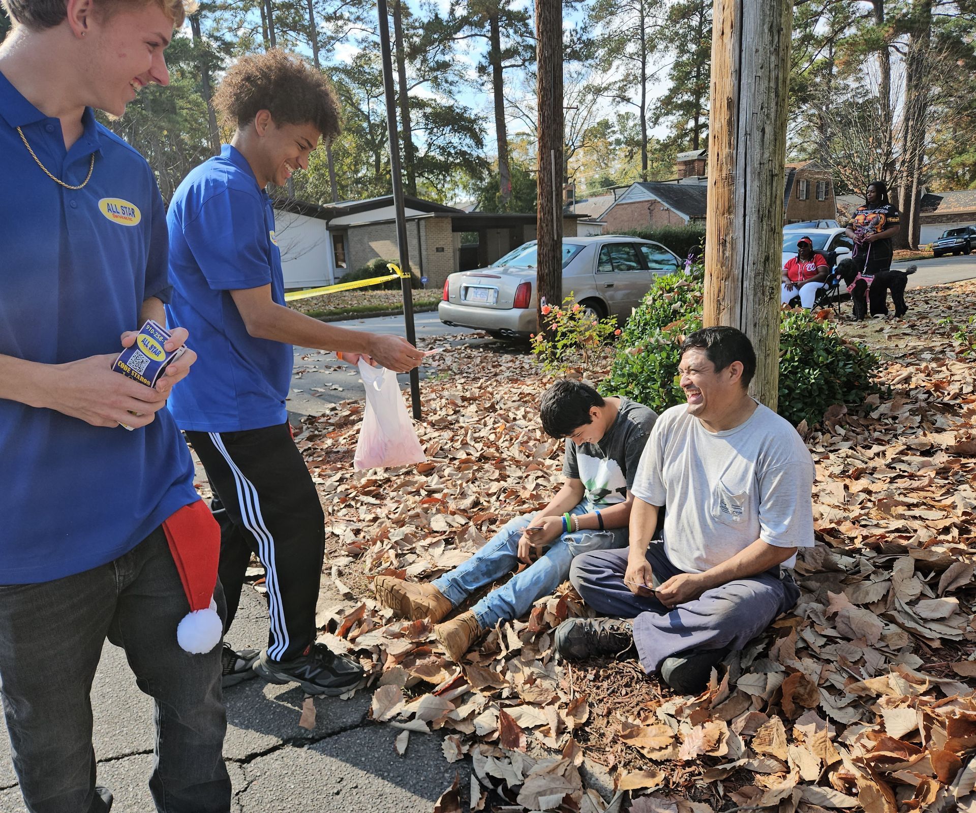 Two young people in blue shirts interact with two others sitting on a leaf-covered lawn near a tree and parked cars.