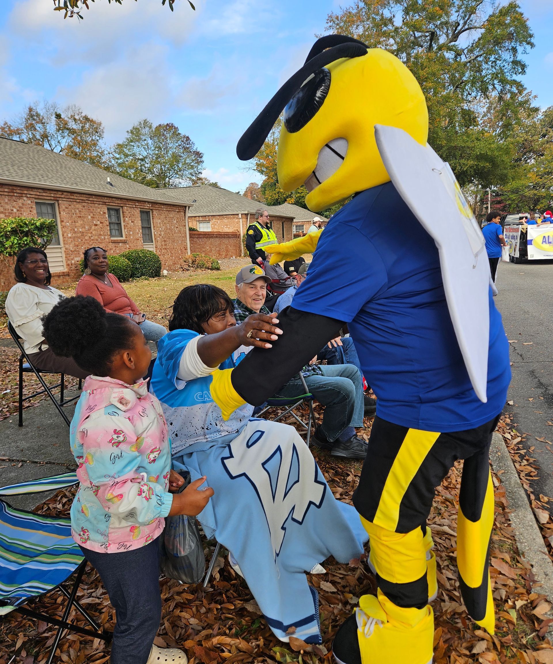A yellow and black bee mascot greets a person wrapped in a blue blanket as a child watches, all sitting outdoors.