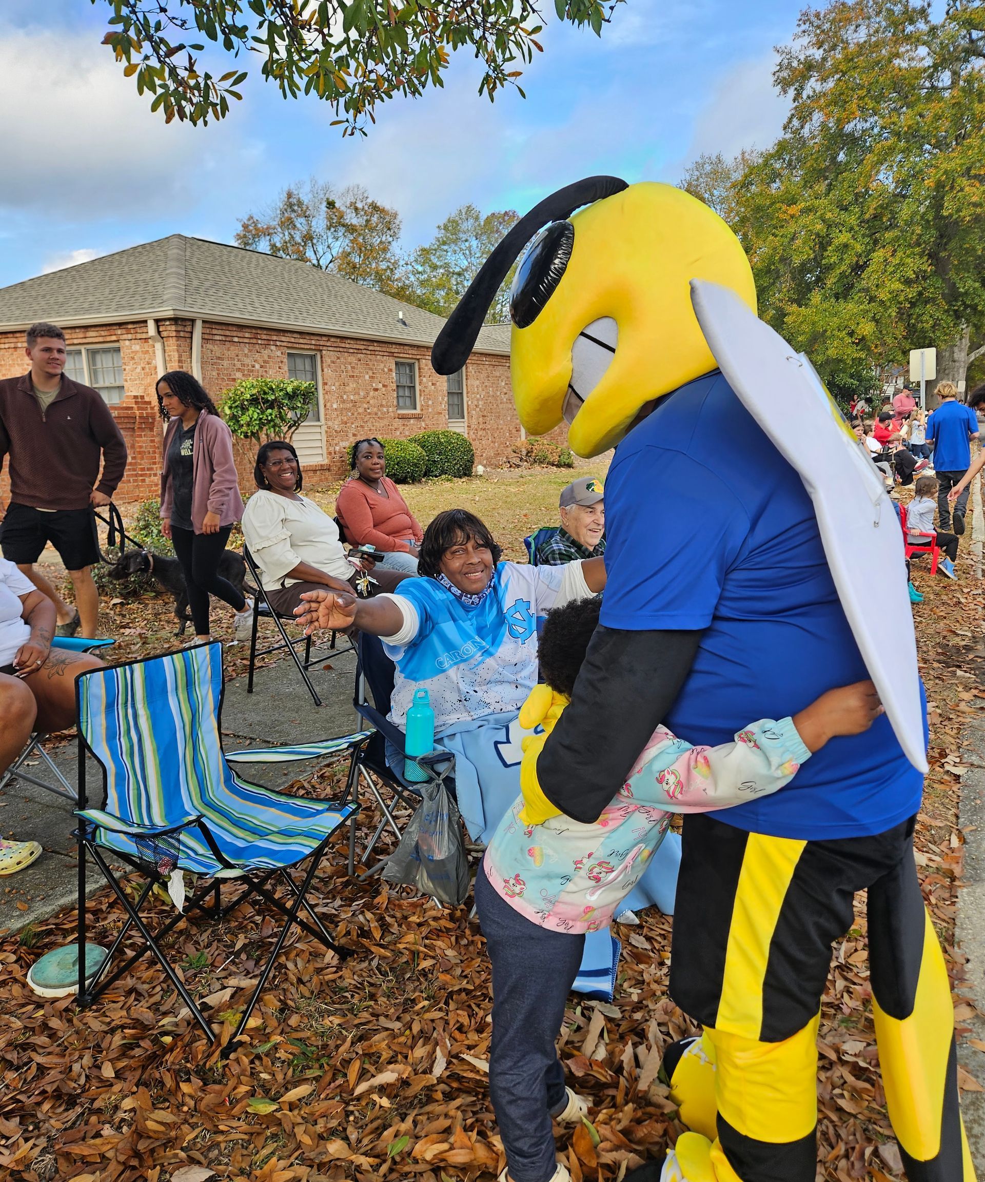 A mascot shaped like a yellow and blue bee hugs a person outdoors, while others watch from folding chairs in autumn.