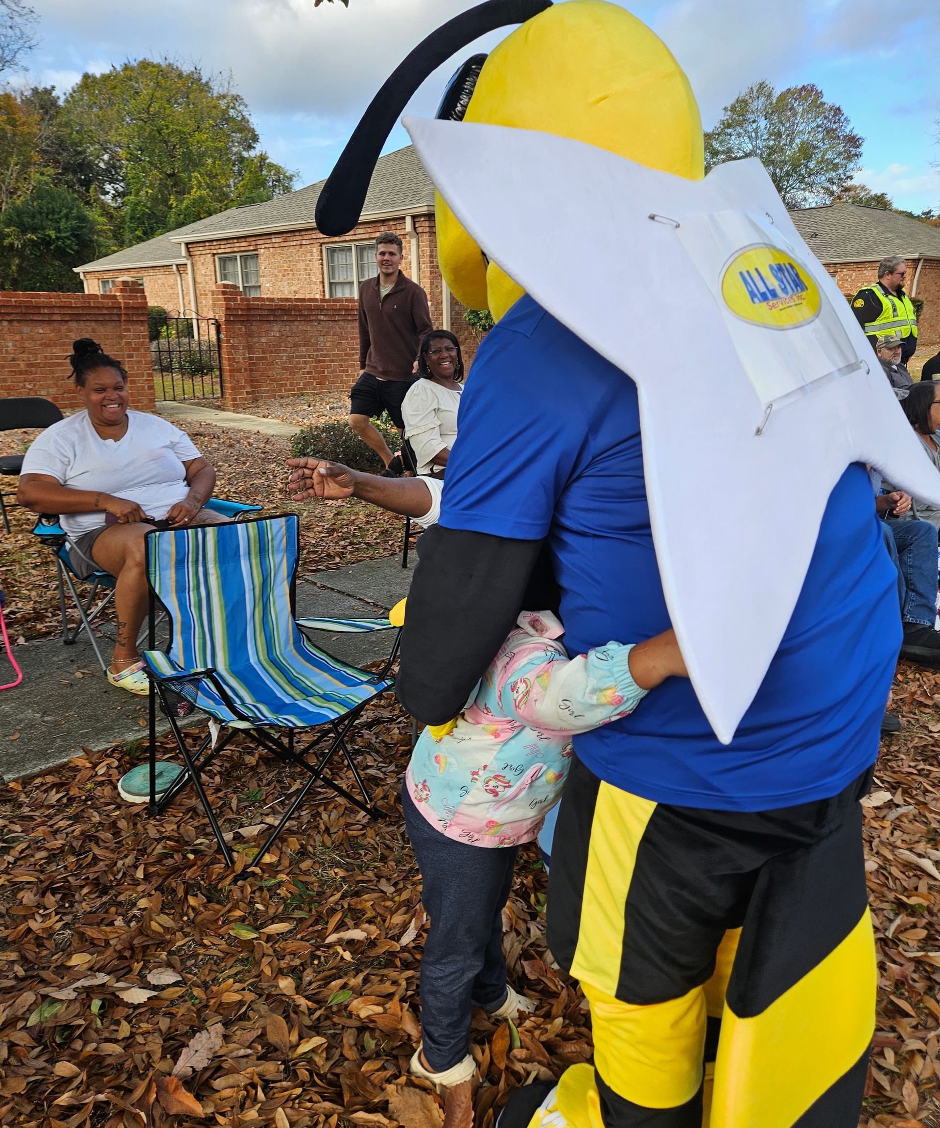 A child in a patterned shirt hugs a mascot dressed as a bumblebee in a yard filled with autumn leaves.