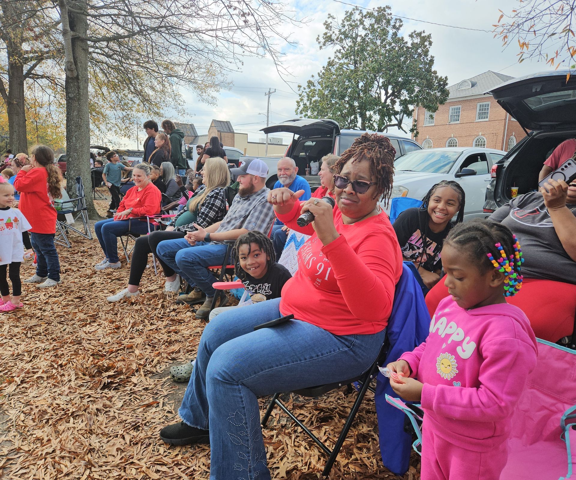 People seated in chairs on a leaf-covered field at an outdoor community event, with some attendees facing the camera.