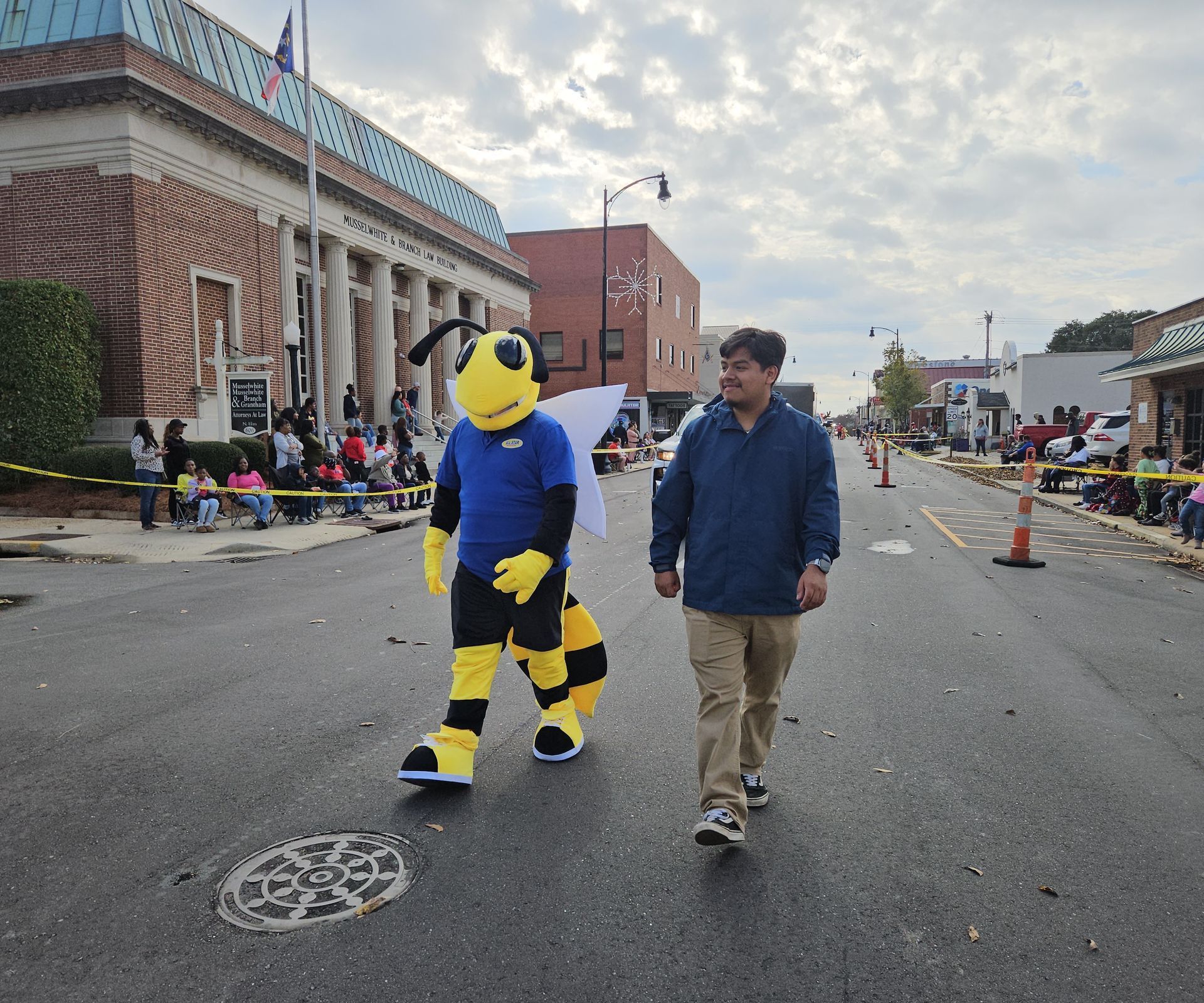 A mascot dressed as a yellow and black bee walks down a street next to a person in a blue jacket.