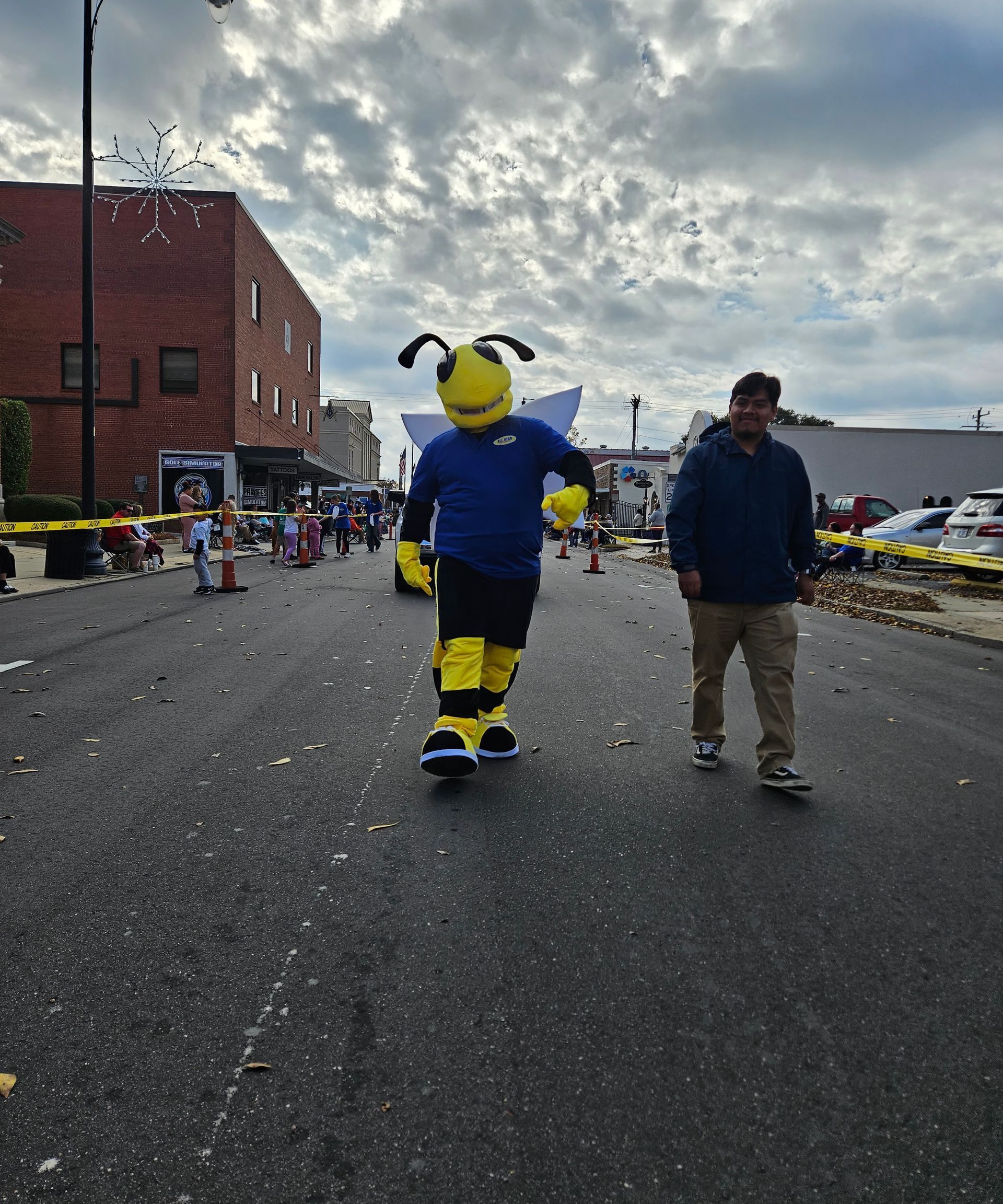 A person in a yellow and blue bumblebee mascot costume walking down a street alongside a person in a blue jacket.