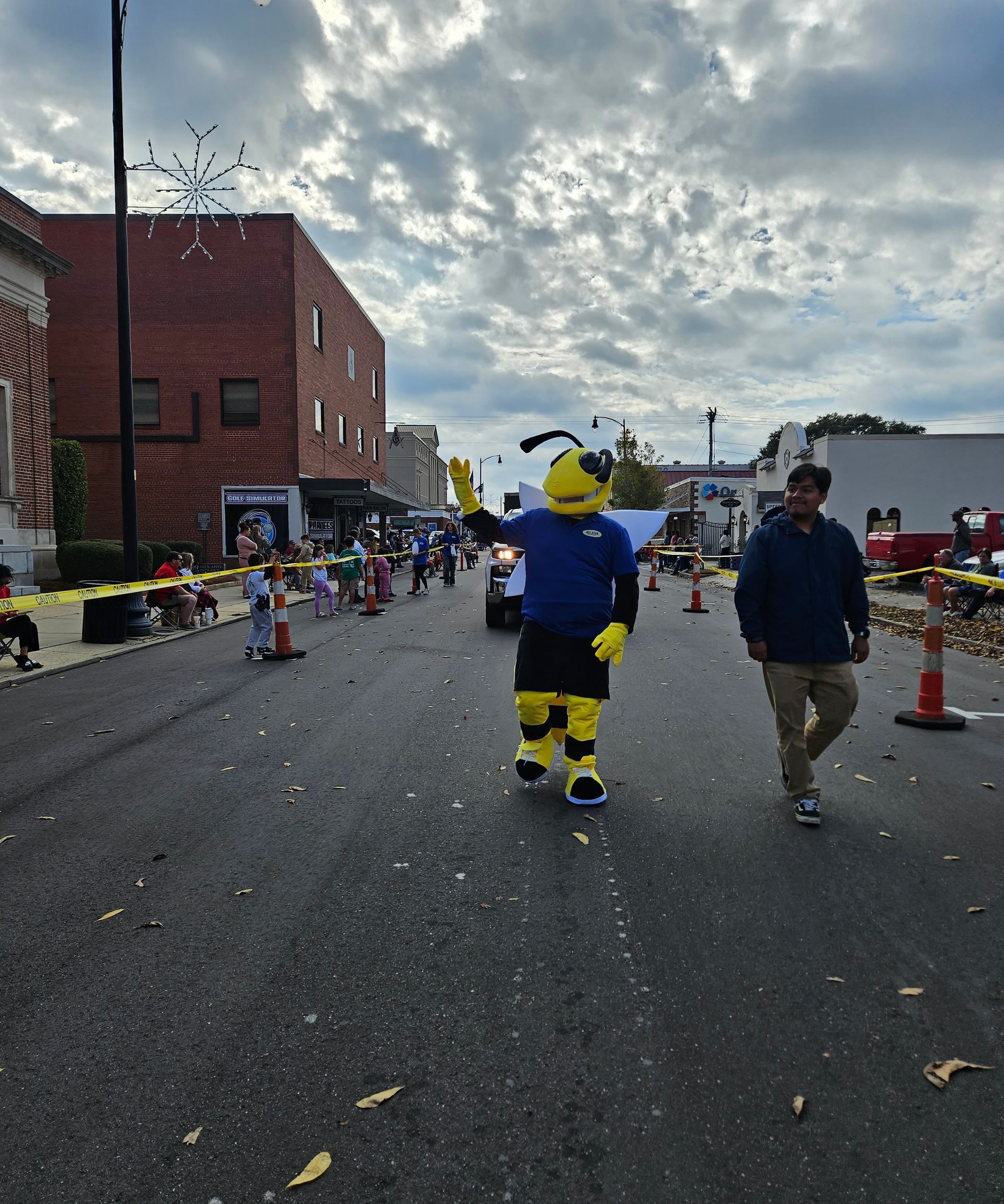 A yellow and black bee mascot waves while walking down a street parade, accompanied by a person in a blue jacket.