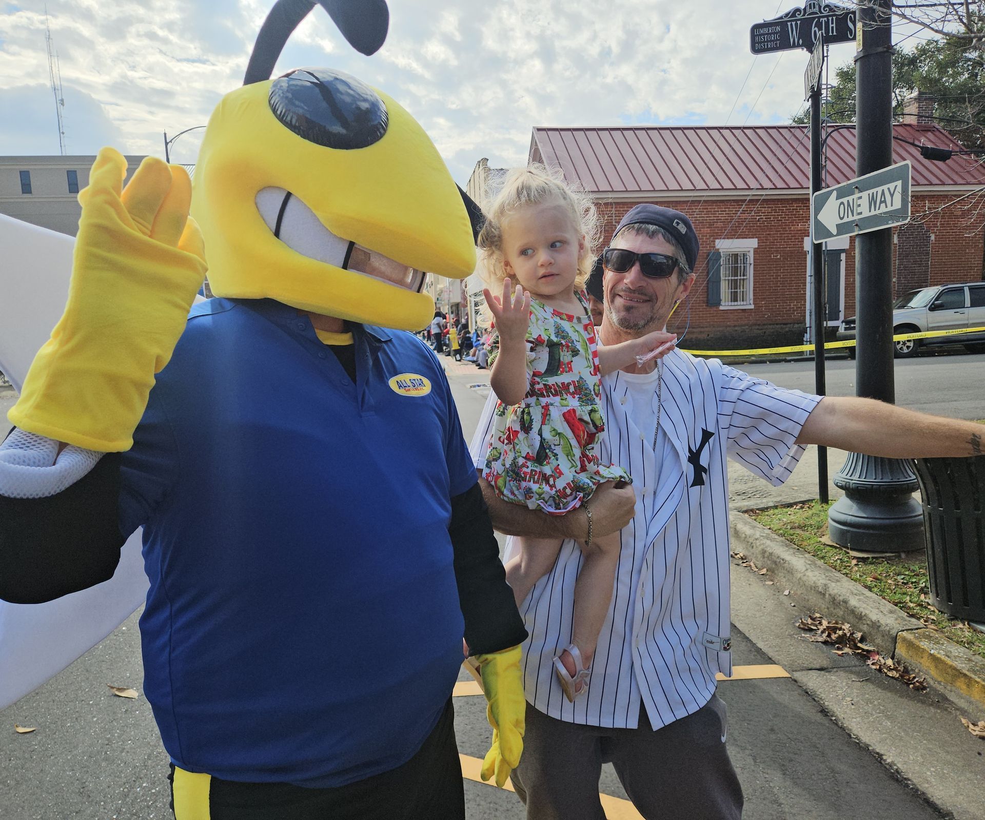 A yellow mascot waves next to a person holding a small child outdoors, with a street and a brick building in the background.
