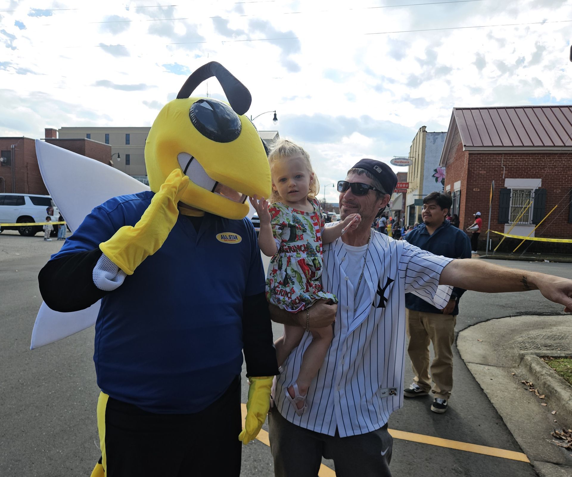 A costumed yellow bee mascot, a person holding a small child, and another person stand together on a sunny town street.