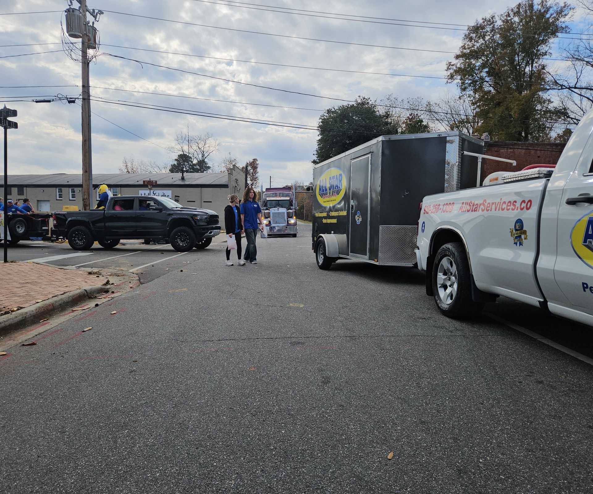 Two people walk across a paved street between a dark pickup truck and a white truck towing a gray trailer.
