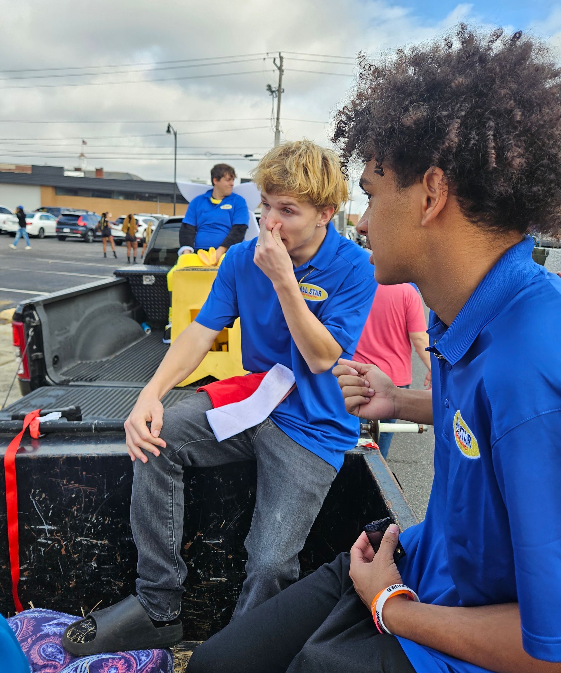 Two people in matching blue polo shirts sit in the back of a truck in a parking lot.