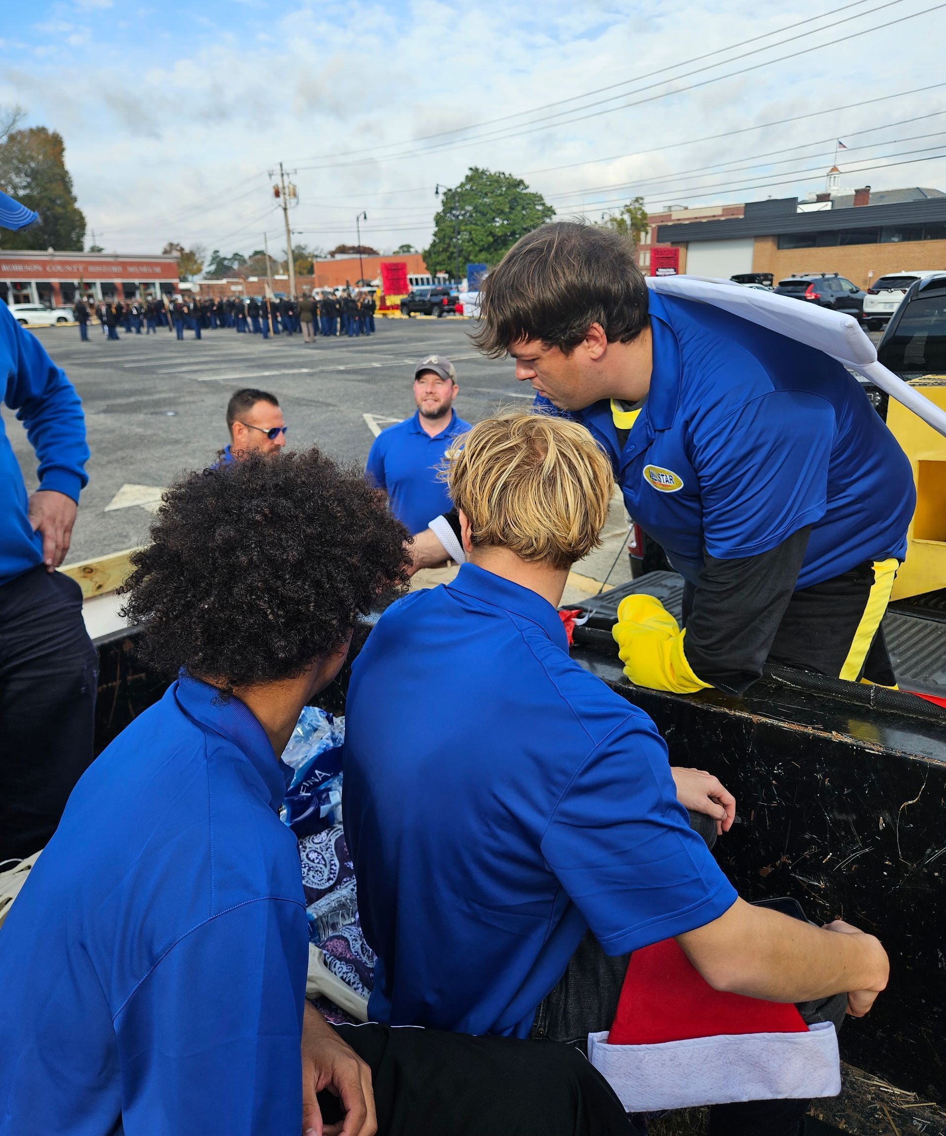 People wearing matching blue shirts decorate a float outdoors near a parking lot with other groups in the background.