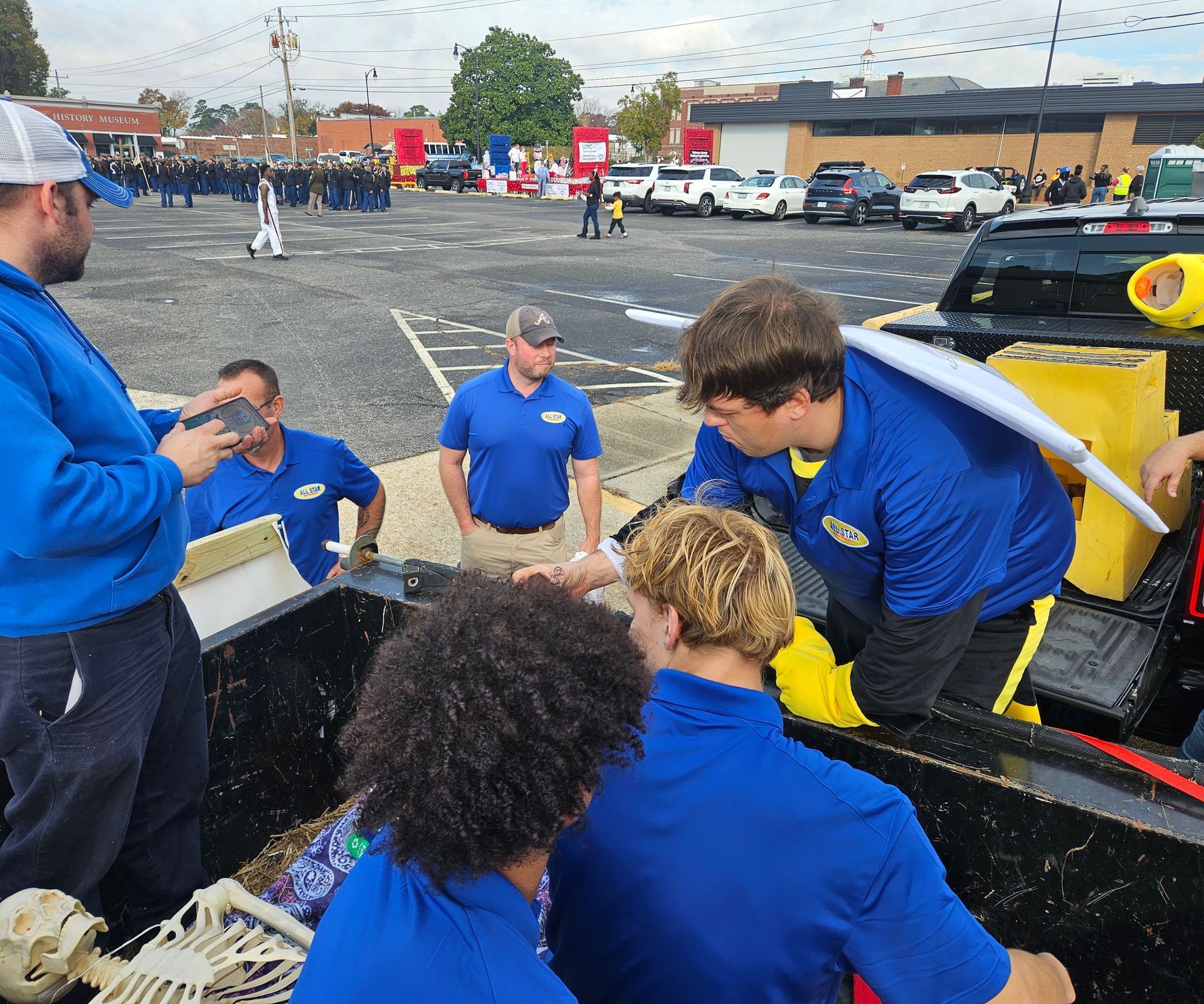 A group of people wearing blue shirts work together in the back of a truck, assembling a skeleton in a parking lot.