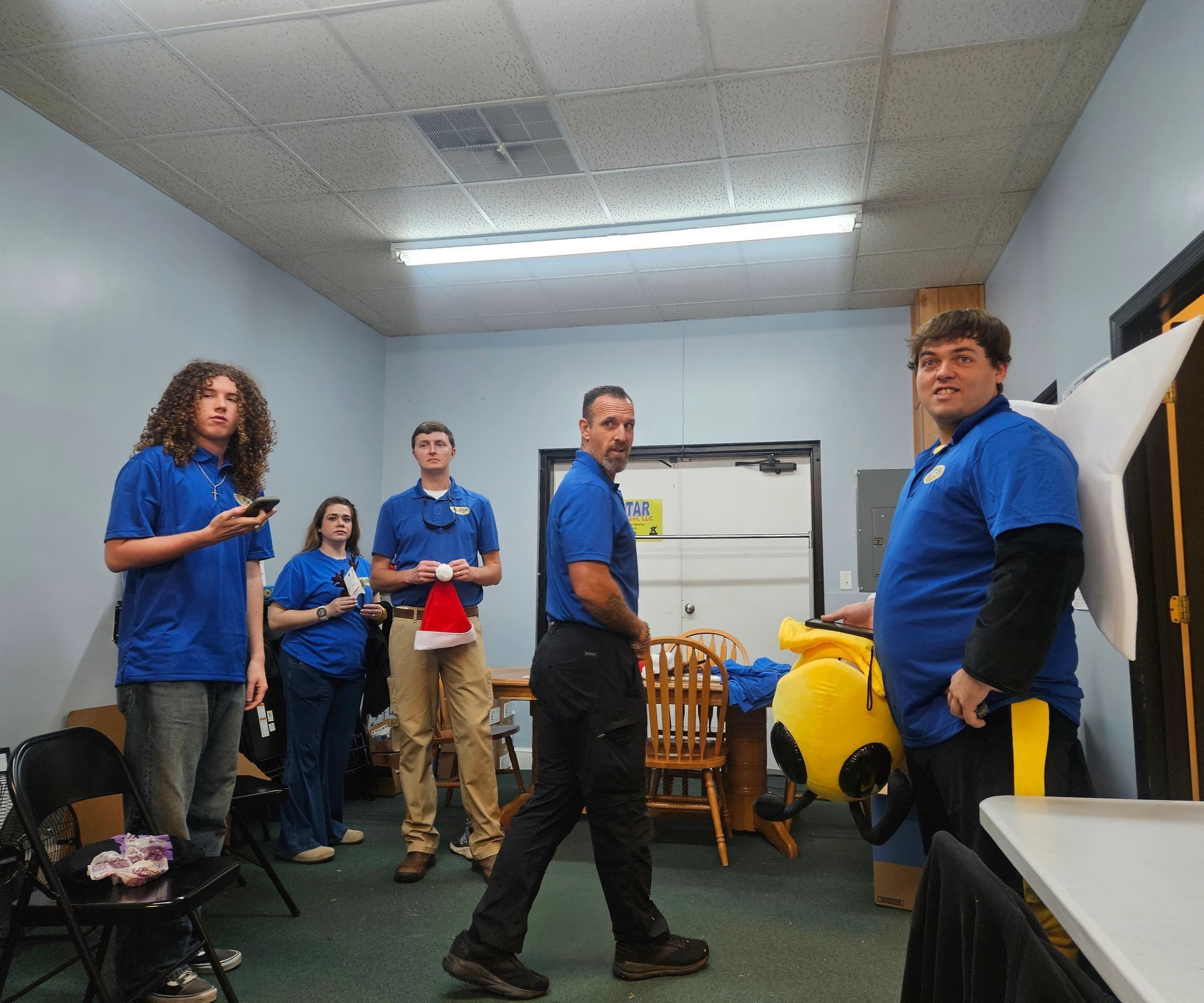 Group of people wearing blue shirts in an office room, one holding a Santa hat and another holding a yellow mascot head.