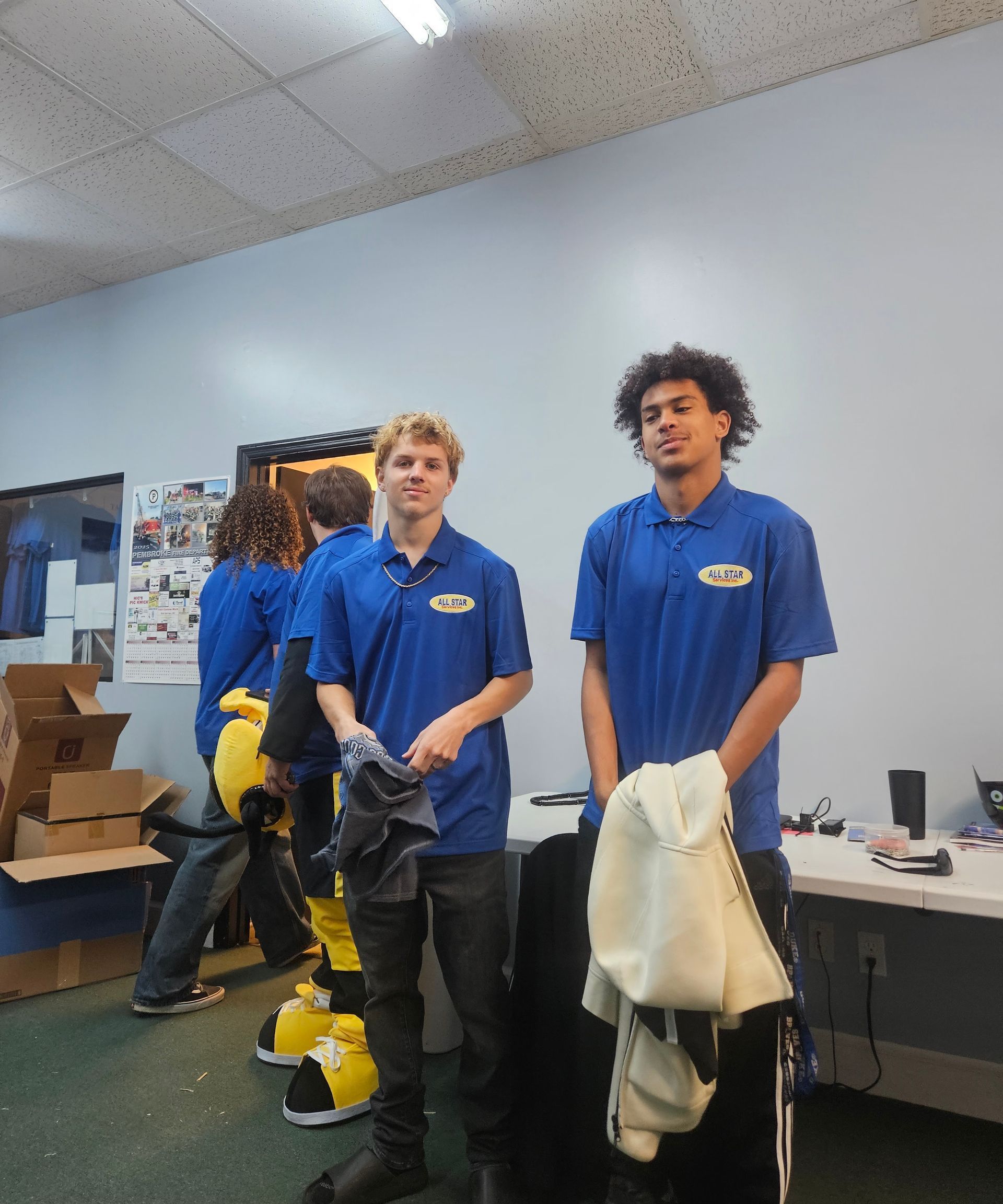 Two young people in blue polo shirts stand in an indoor office space with a yellow mascot costume nearby.